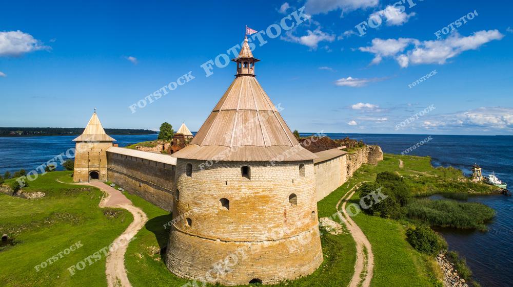 На фотографии изображена крепость Орешек, находящаяся на острове в устье реки Невы, на Ладожском озере. Основанная в XIV веке, крепость была важным оборонительным сооружением и сохранила свои монументальные башни и стены до наших дней. Солнечный день подчеркивает текстуру каменной кладки и историческое значение этого места. Окружающая природа Ладожского озера добавляет пейзажу живописности и покоя.